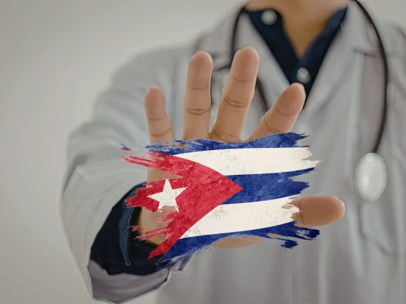 Close up of a doctor with a stethoscope holding out his hand in a “Stop“ motion as he grabs shards of a Cuban flag.